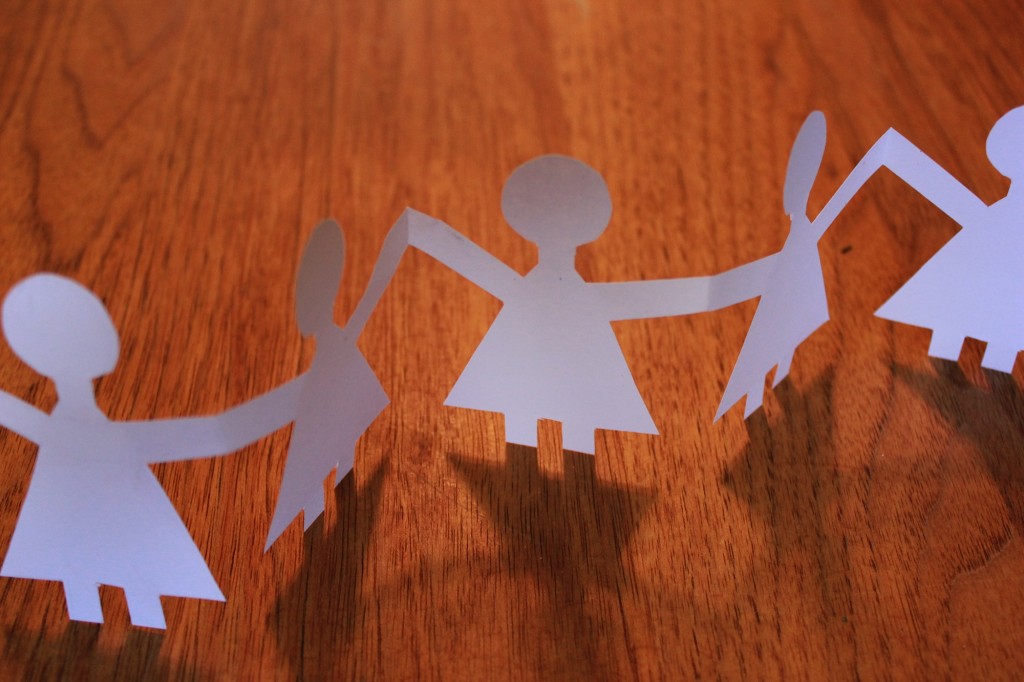 Chain of paper cutout figures holding hands on a wooden table surface, white and brown tones, cheerful crafted mood.