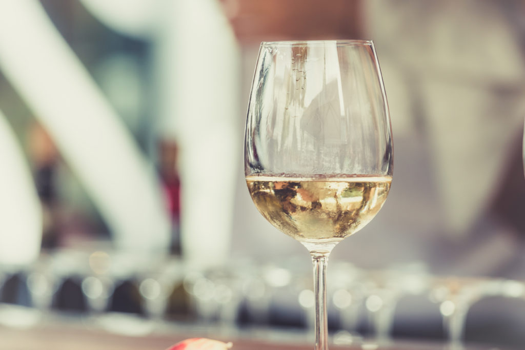 Close-up of a wine glass with pale golden liquid, blurred bright background, elegant and warm mood.